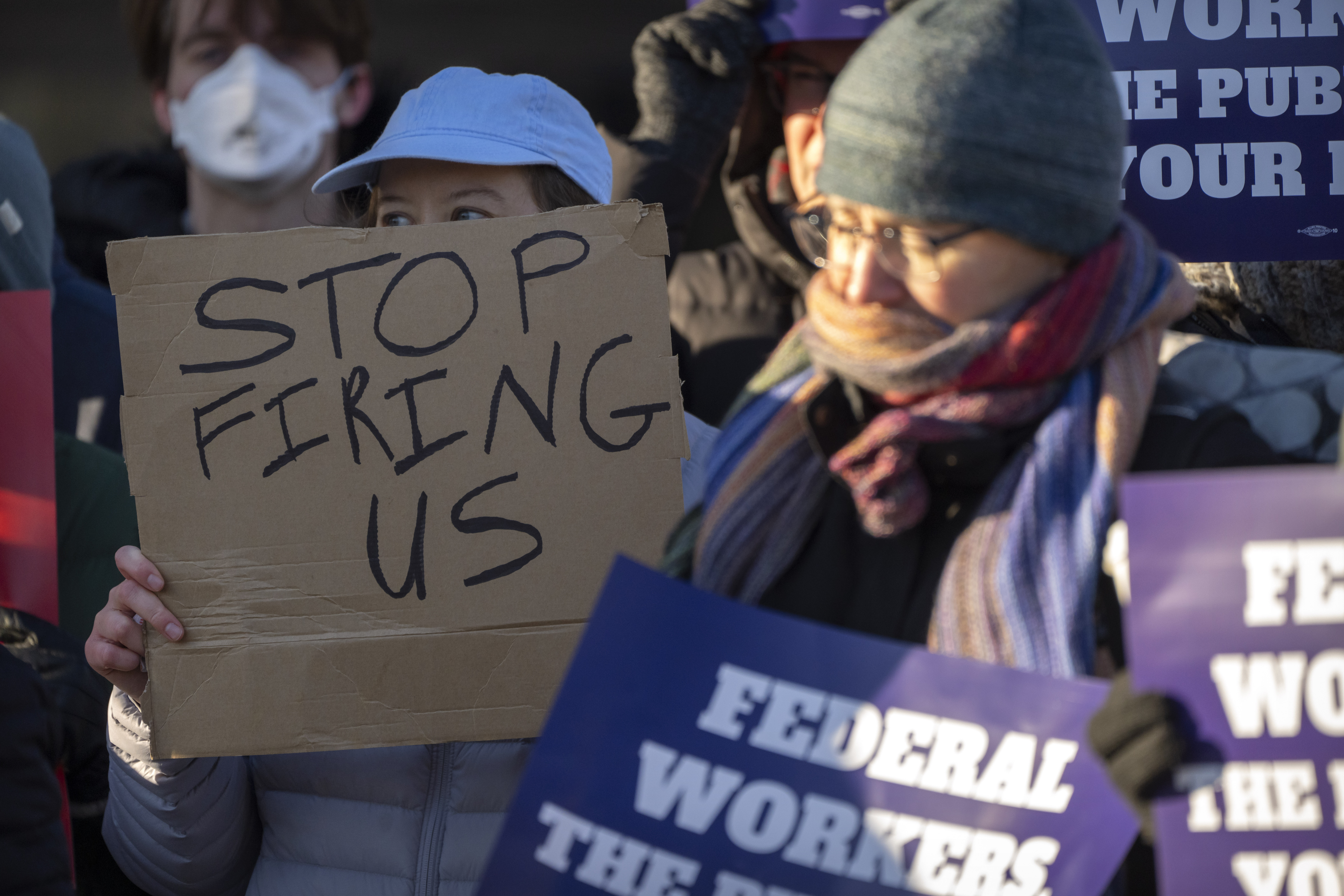 Demonstrators rally, and one holds a sign that says "Stop firing us." Demonstrators rally, and one holds a sign that says "Stop firing us."